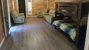 A room with wooden walls and a sloped ceiling, featuring a bunk bed, two leather chairs, and a ceiling fan.