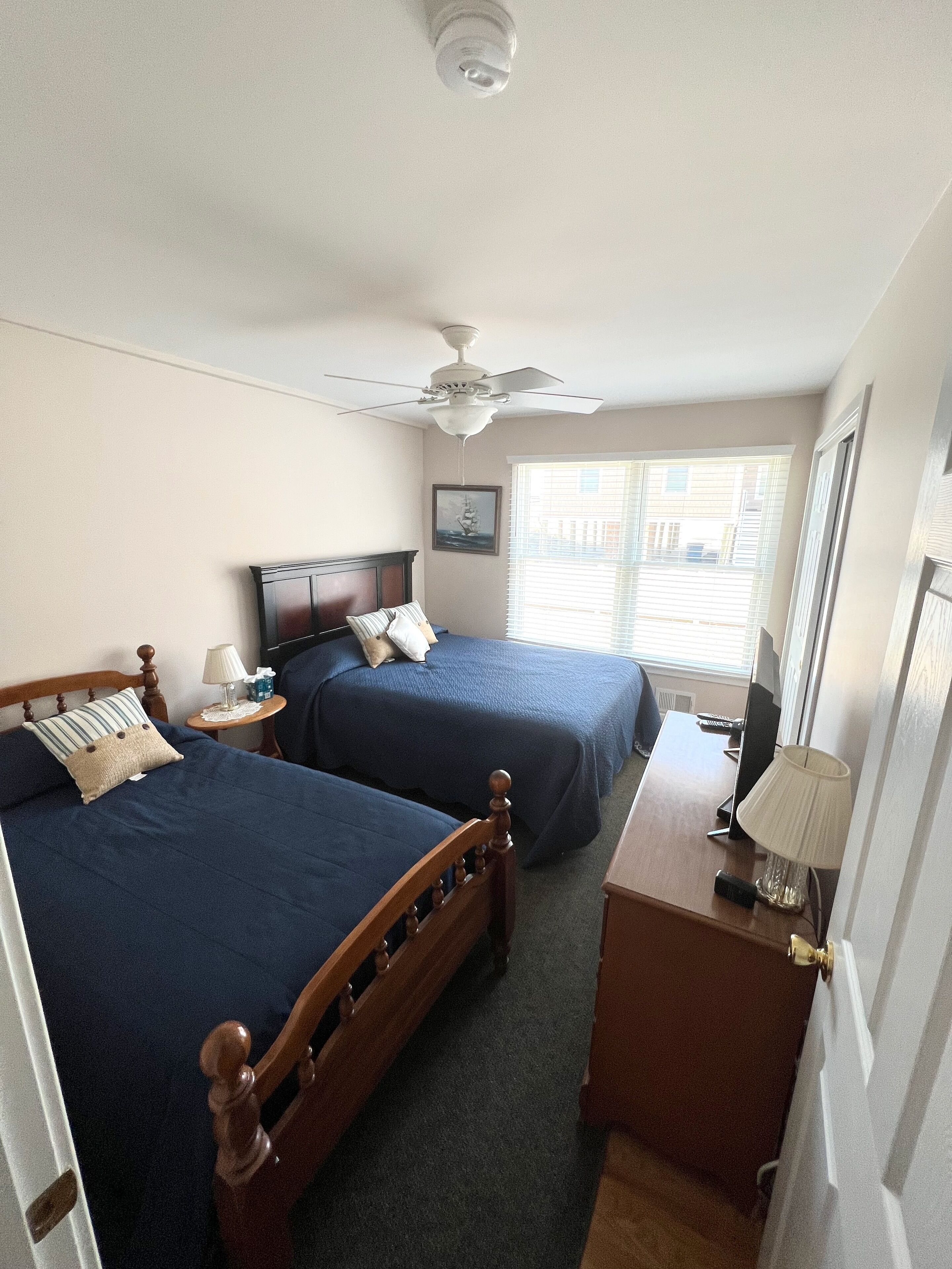 A bedroom with two beds, a ceiling fan, a bedside table with a lamp, and a window with blinds.