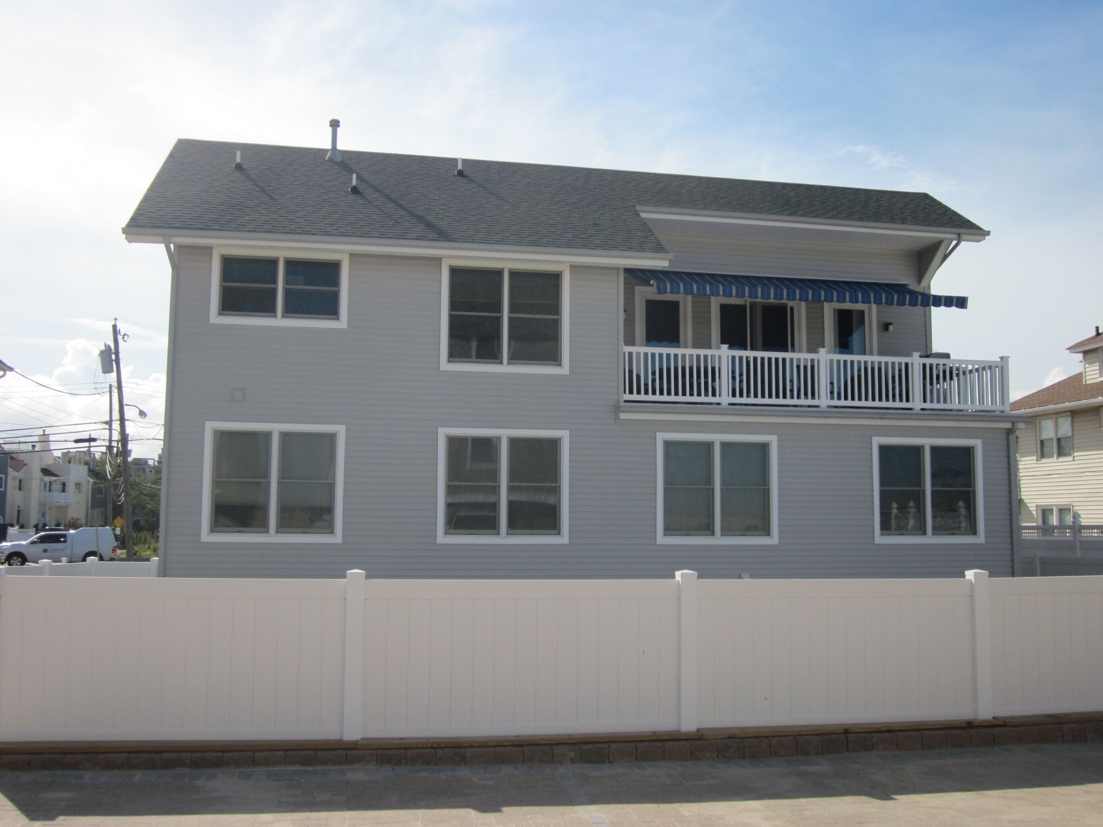 A two-story residential building with a balcony on the second floor, a white fence in front, and a clear sky.