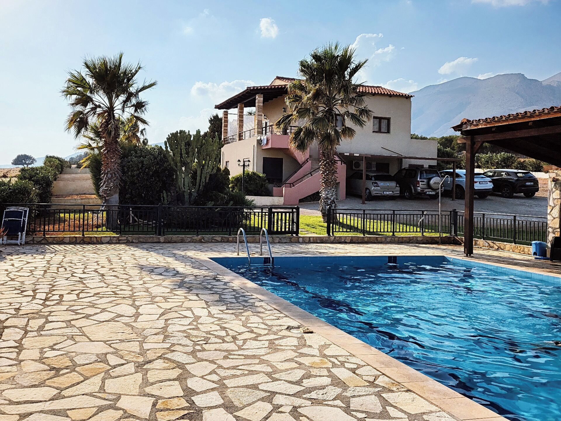 A swimming pool with stone tiles, a building with a pink facade, and parked cars in the background.