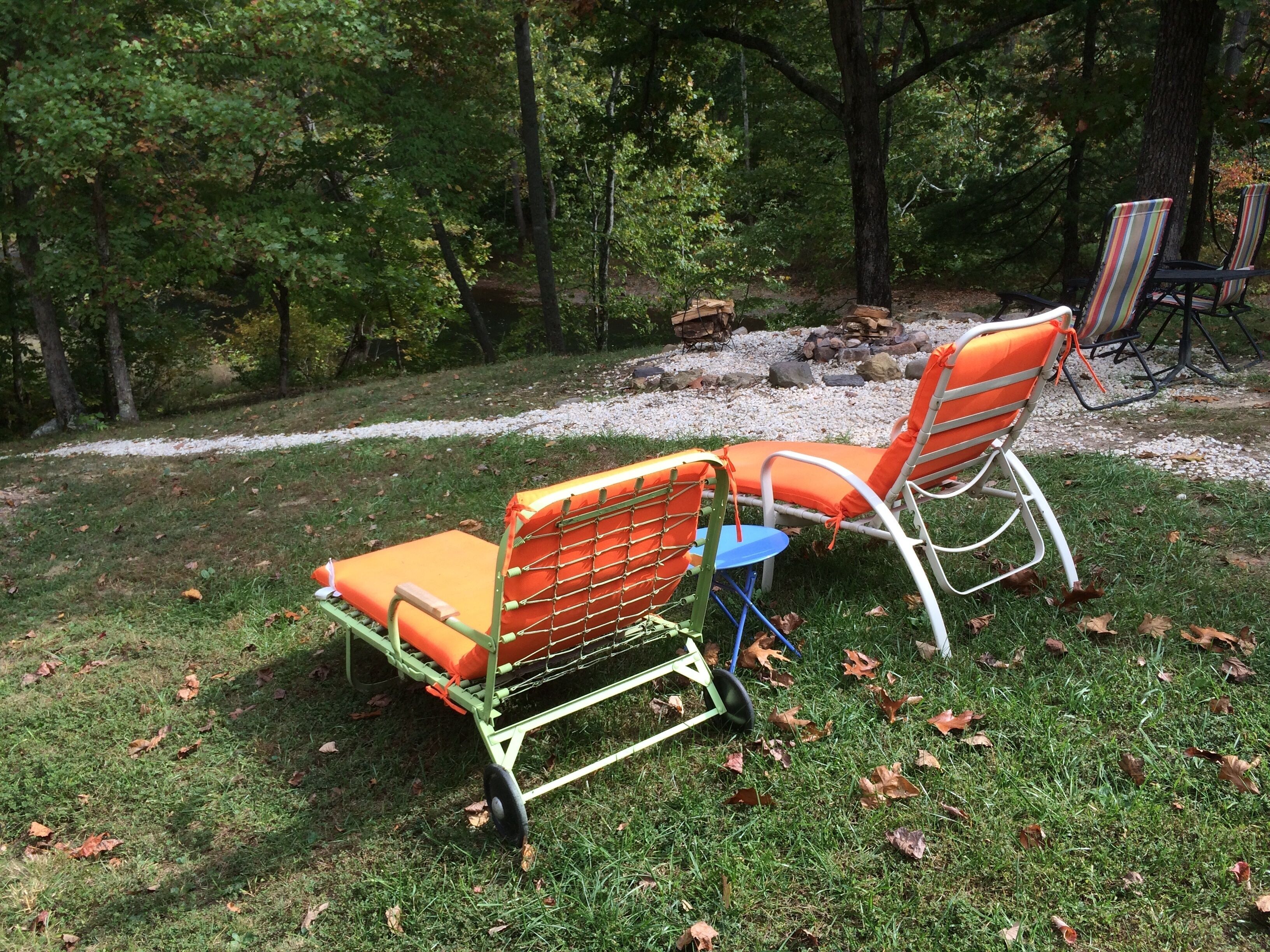 Zwei Gartenstühle mit orangen Kissen und ein kleiner Tisch, auf einer grünen Wiese vor Bäumen.
