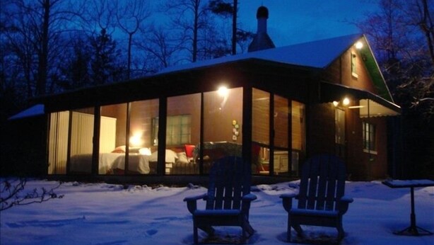 Een chalet met grote ramen, twee Adirondack-stoelen buiten en een met sneeuw bedekte grond.