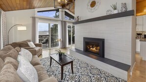 A modern living room with a fireplace, a large sofa, and a dining area visible through a glass door. - Pleasant Beach Getaway - Breathtaking Puget Sound Views and Steps from the Beach (Bainbridge Island)