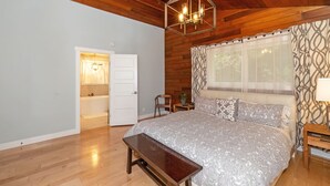 A bedroom with a wooden ceiling, a large bed with patterned bedding, a bench, a bedside table, and a window with patterned curtains. - Pleasant Beach Getaway - Breathtaking Puget Sound Views and Steps from the Beach (Bainbridge Island)