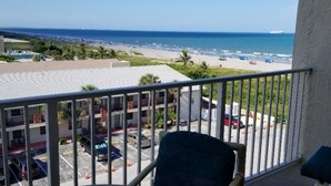 A balcony with a view of a beach and ocean. There are parked cars and a building with multiple floors in the background.