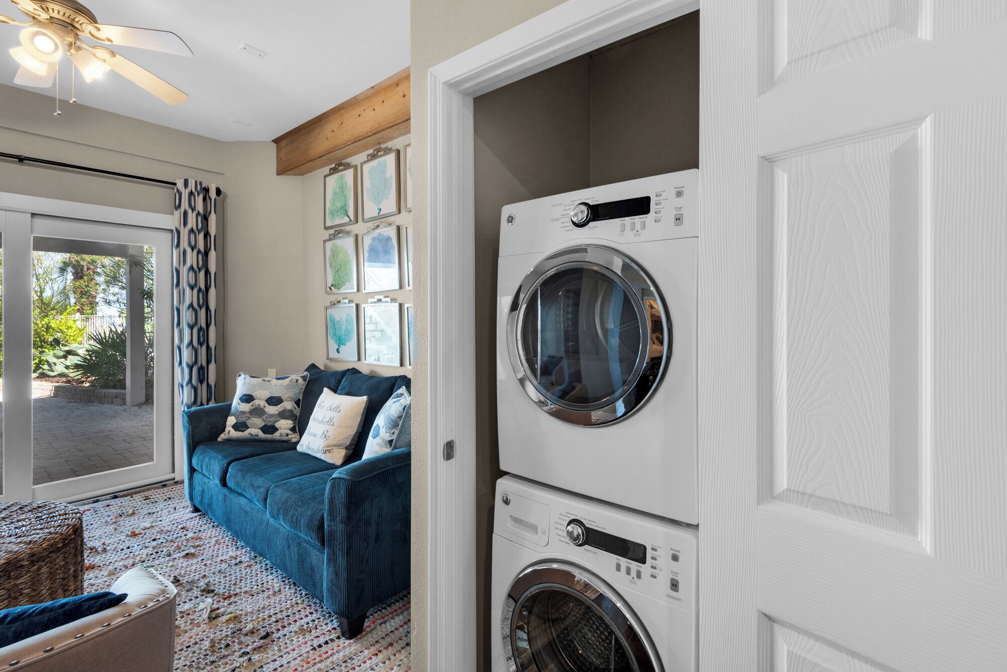 A laundry room with a front-loading washer and dryer, a blue sofa with patterned cushions, and a view of a patio with a ceiling fan.