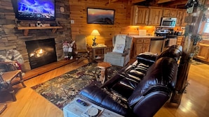Ein gemütliches Blockhaus-Interieur mit Kamin aus Stein, Ledersessel, Holzesstisch und an der Wand montiertem Fernseher.