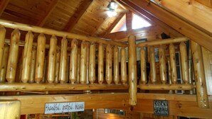 Ein Holzblockhaus im Inneren mit einem Erkerbereich, der sich durch eine Holzrelingung auszeichnet. Auf den Wänden stehen Schilder mit den Aufschriften 'thankful, grateful, blessed' und 'If you are lucky enough to be at the cabin'.