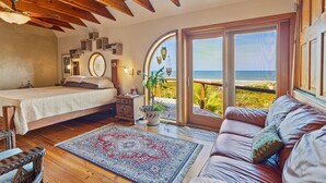Une chambre à coucher avec un lit, un plafond en bois, une grande fenêtre donnant sur la mer, un canapé en cuir marron et un tapis à motifs.