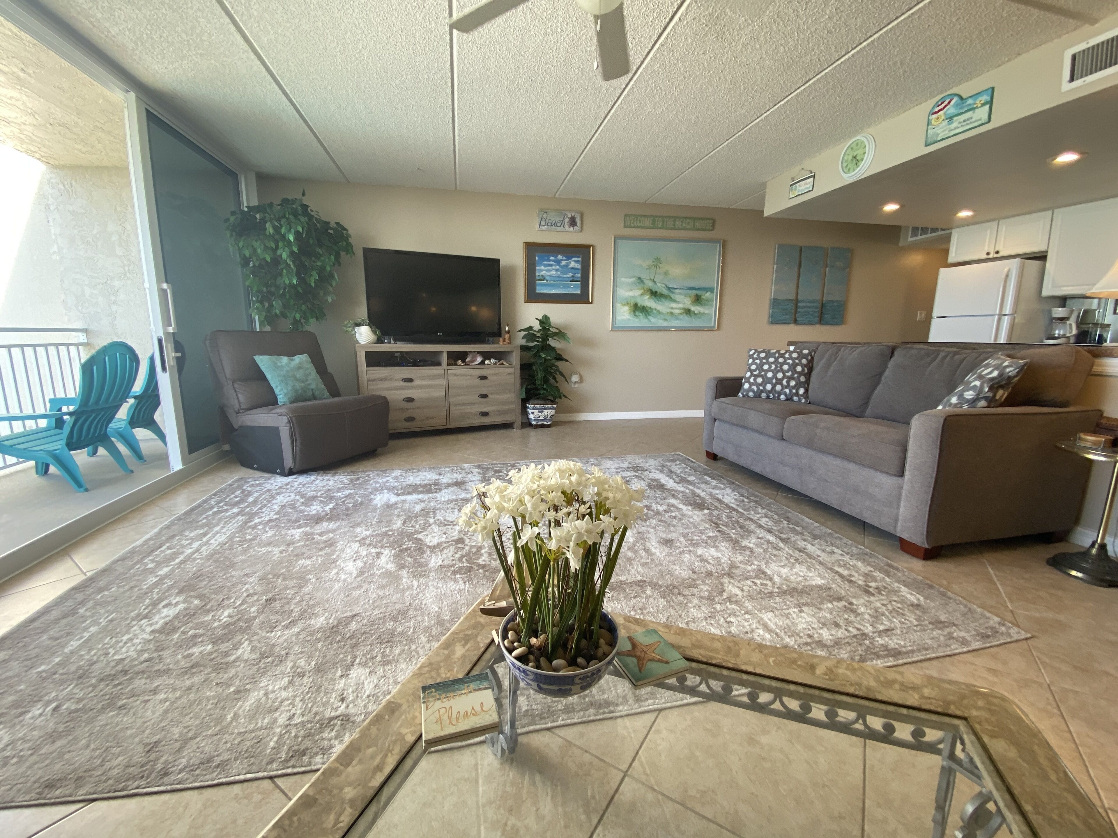 A living room with a flat-screen TV, a gray sofa, a gray armchair, a coffee table with a vase of flowers, and a large window.