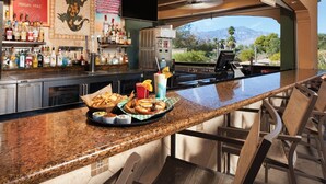 A bar area with a granite countertop, bar stools, a variety of bottles, a menu board, and a view of mountains.