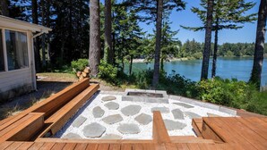 Una terraza de madera con escalones de piedra que conducen a una zona con brasero, rodeada de árboles altos y vistas a un lago.