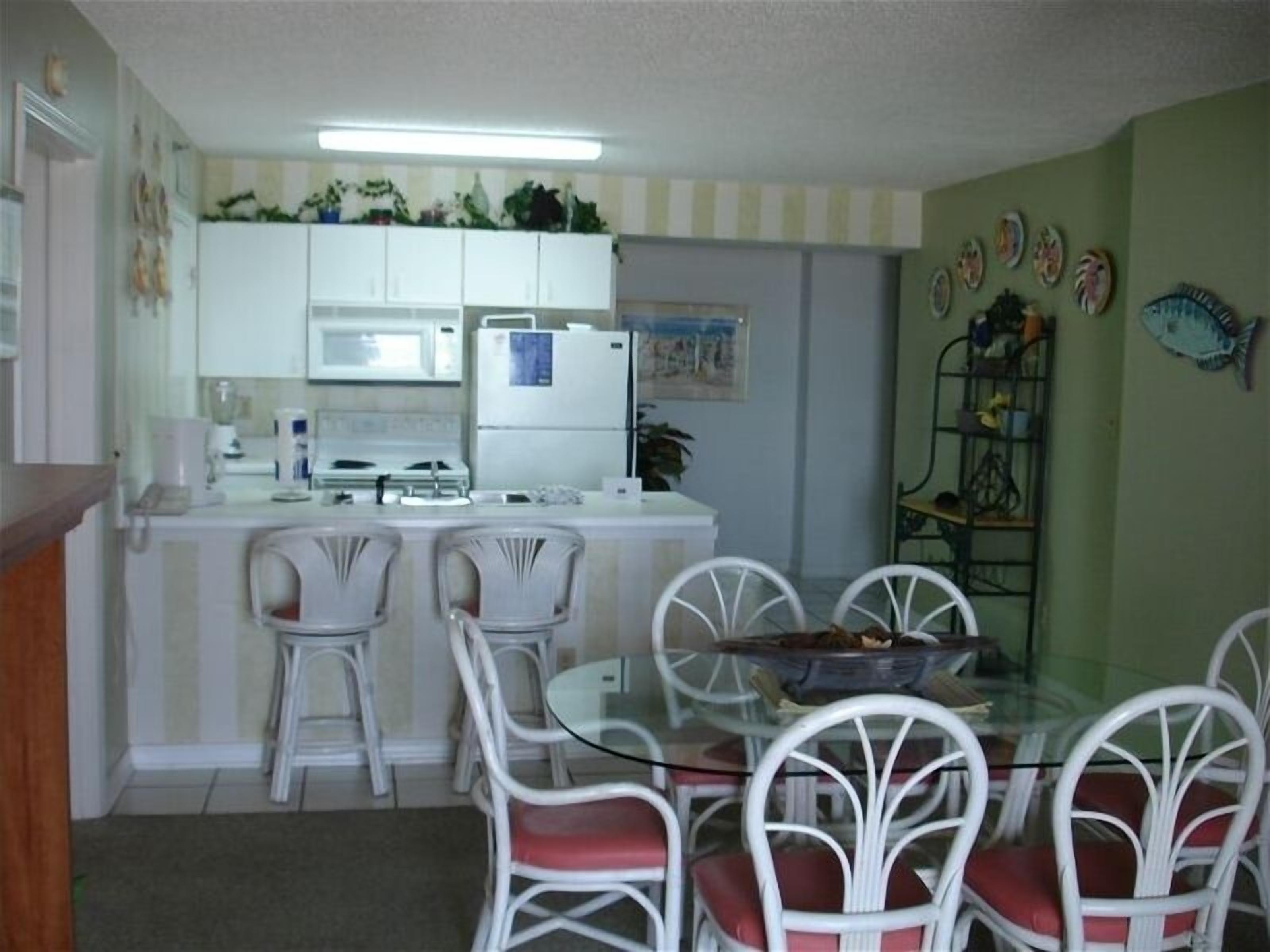 A compact kitchen with white cabinets, a microwave, a refrigerator, and a stove. There are four white chairs with red cushions around a glass table.