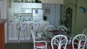 A compact kitchen with white cabinets, a microwave, a refrigerator, and a stove. There are four white chairs with red cushions around a glass table. - Relax! Put Your toes in the Sand at this DIRECT GULF FRONT CONDO   (Orange Beach)