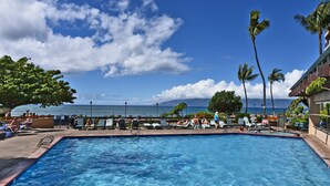 Una piscina con agua azul cristalina, rodeada de sillones de descanso y palmeras, con vista a una playa y al océano.