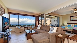 Amplia sala de estar con un ventanal que ofrece vistas al mar y edificios, un televisor de pantalla plana y un rincón de descanso acogedor con un sofá y sillones.