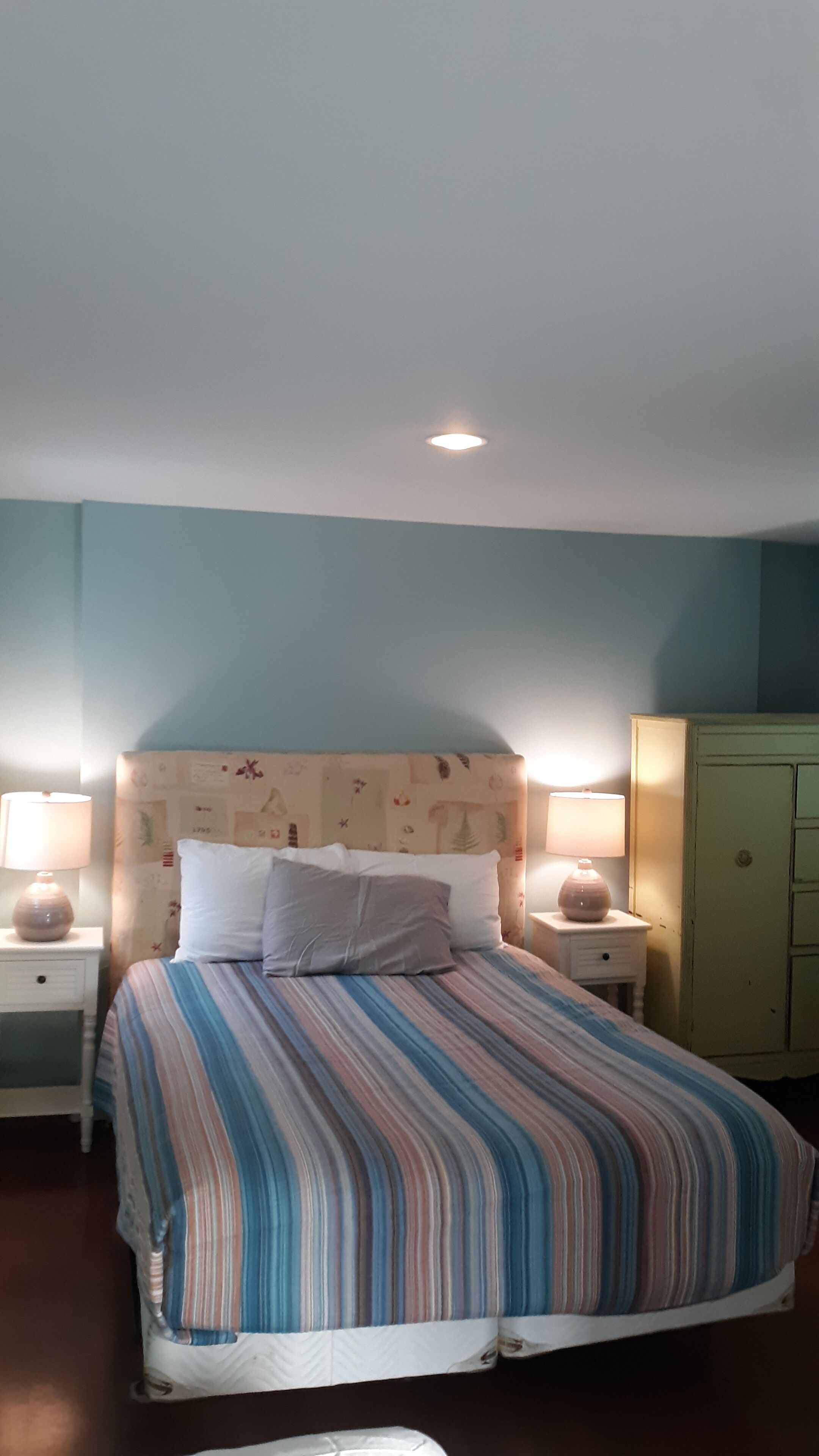 Een net opgemaakt bed met streepjesbeddengoed, twee bedlampjes, een hoofdbord met een bloemenprint en een klein kastje aan de rechterkant.
