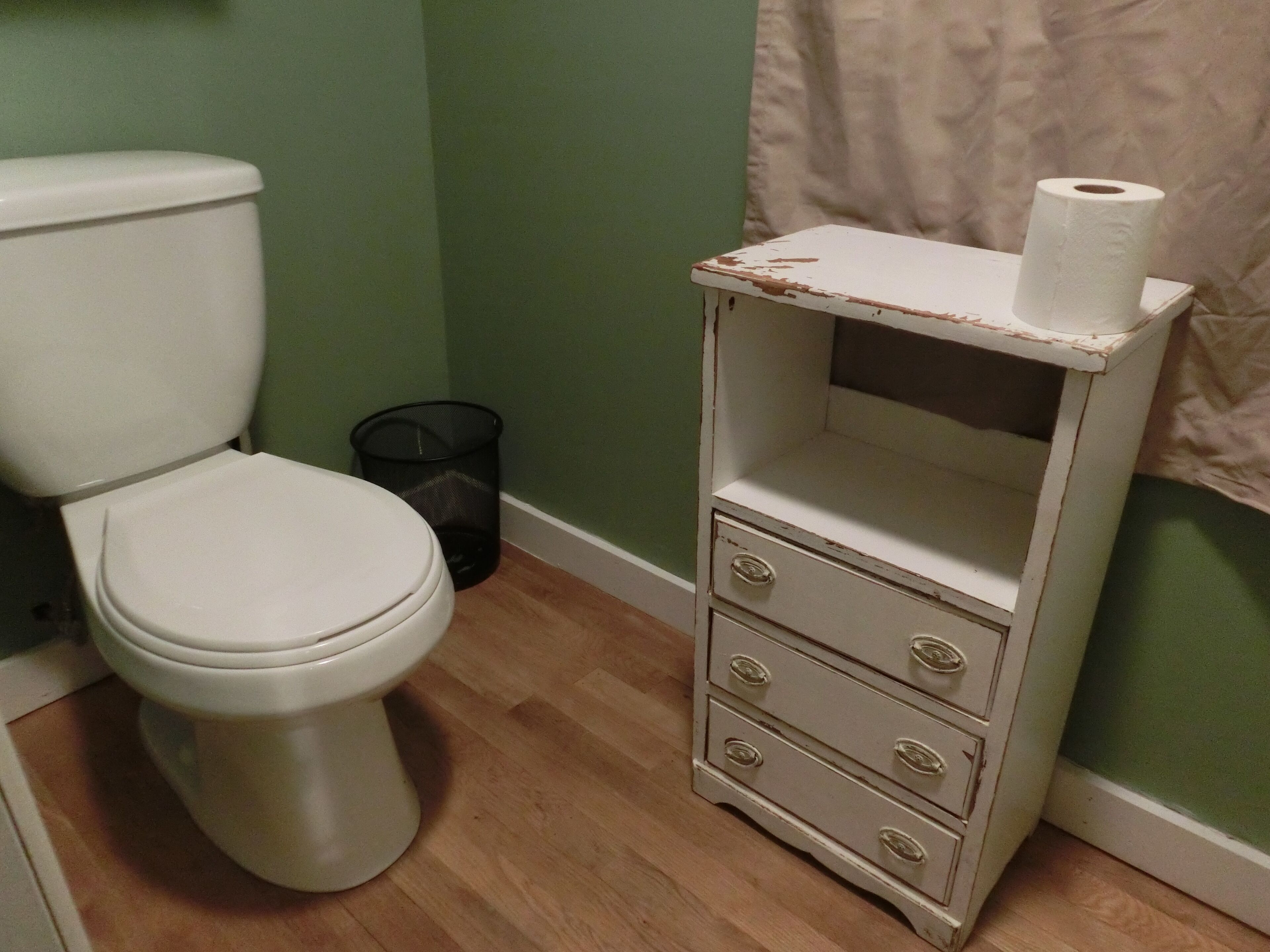 Un baño con inodoro, un pequeño cubo de basura negro y un aparador blanco con cajones.