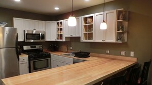A modern kitchen with white cabinets, a stainless steel refrigerator, a microwave, and a stove. There are pendant lights above the island and a sink with a black faucet. - Quaint,,,Quiet..up island feel yet a short walk to town and harbor (Oak Bluffs)
