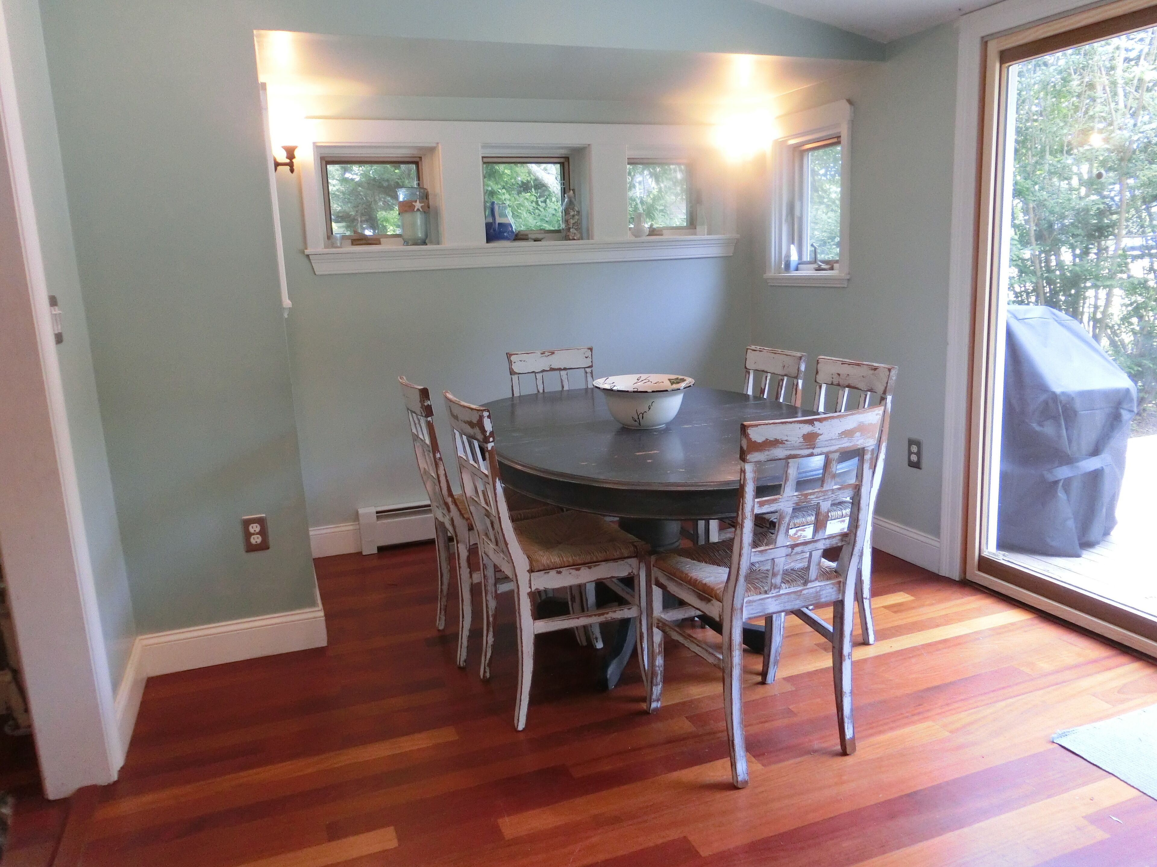 A dining room with a round table surrounded by six chairs, a window with a view of trees, and a covered grill outside.