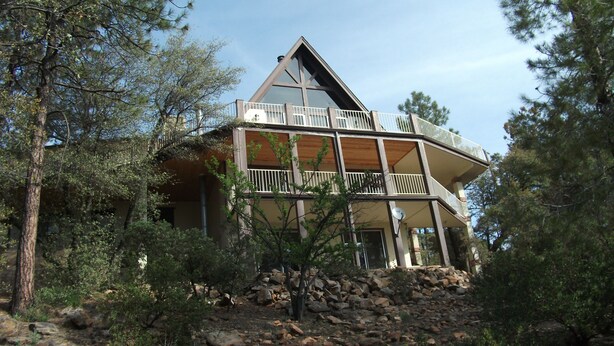A multi-story house with a prominent triangular roof, surrounded by trees and a rocky landscape.