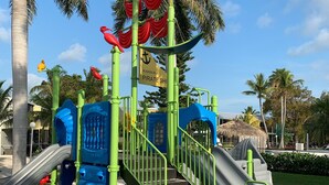 A playground with slides and climbing structures, surrounded by palm trees and a clear sky. - Updated Key Largo Townhome • Pool • Saltwater Lagoon • Beach • Oceanfront Access (Key Largo)