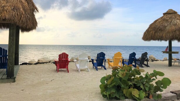 A beach setting with thatched umbrellas, a row of colorful Adirondack chairs, and a sandy beach. - Updated Key Largo Townhome • Pool • Saltwater Lagoon • Beach • Oceanfront Access (Key Largo)