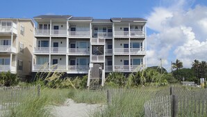 Un immeuble résidentiel à plusieurs étages, doté de balcons, d’un chemin bordé de sable et entouré de végétation.