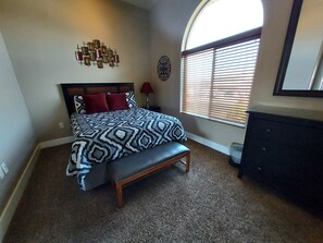 A bedroom with a patterned bedspread, a bench, a wall art piece, and a window with blinds.