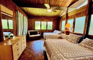 Une chambre de style cabane avec des murs et des planchers en bois, un ventilateur de plafond, deux lits avec une literie à motifs, un petit canapé, une table de chevet avec une lampe et une commode avec des tiroirs.