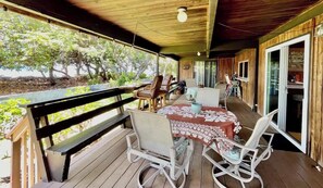 Une terrasse extérieure couverte avec un plancher en bois, une table recouverte d’une nappe à motifs et plusieurs chaises.