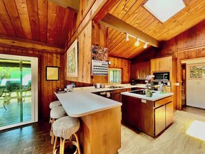 A wooden cabin interior with a kitchen island, bar stools, and a dining area with a view of the outdoors. - Hale Kahakai ~ 2 BR ~ Ocean Front - Puako (Waimea)