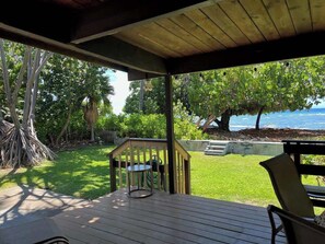 Um deck externo coberto com mesa e cadeiras, com vista para uma área gramada e um corpo d'água.