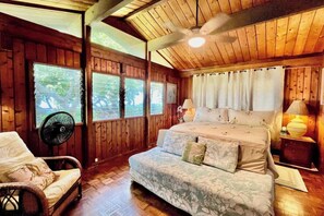 Quarto de cabana de madeira com cama, uma cadeira, um ventilador e uma janela.