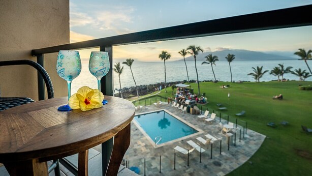 A balcony with a view of a pool and palm trees, a wooden table with a flower, and two glassware. - 407 - Gorgeous Oceanfront one bedroom unit! (Kihei)