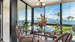 A balcony with a glass table and chairs, a flower vase, and a view of the ocean and palm trees. - 407 - Gorgeous Oceanfront one bedroom unit! (Kihei)