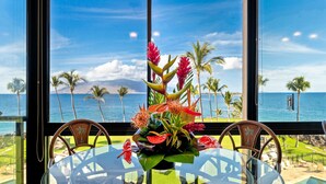 A table with a tropical flower arrangement and a view of the ocean and palm trees. - 407 - Gorgeous Oceanfront one bedroom unit! (Kihei)
