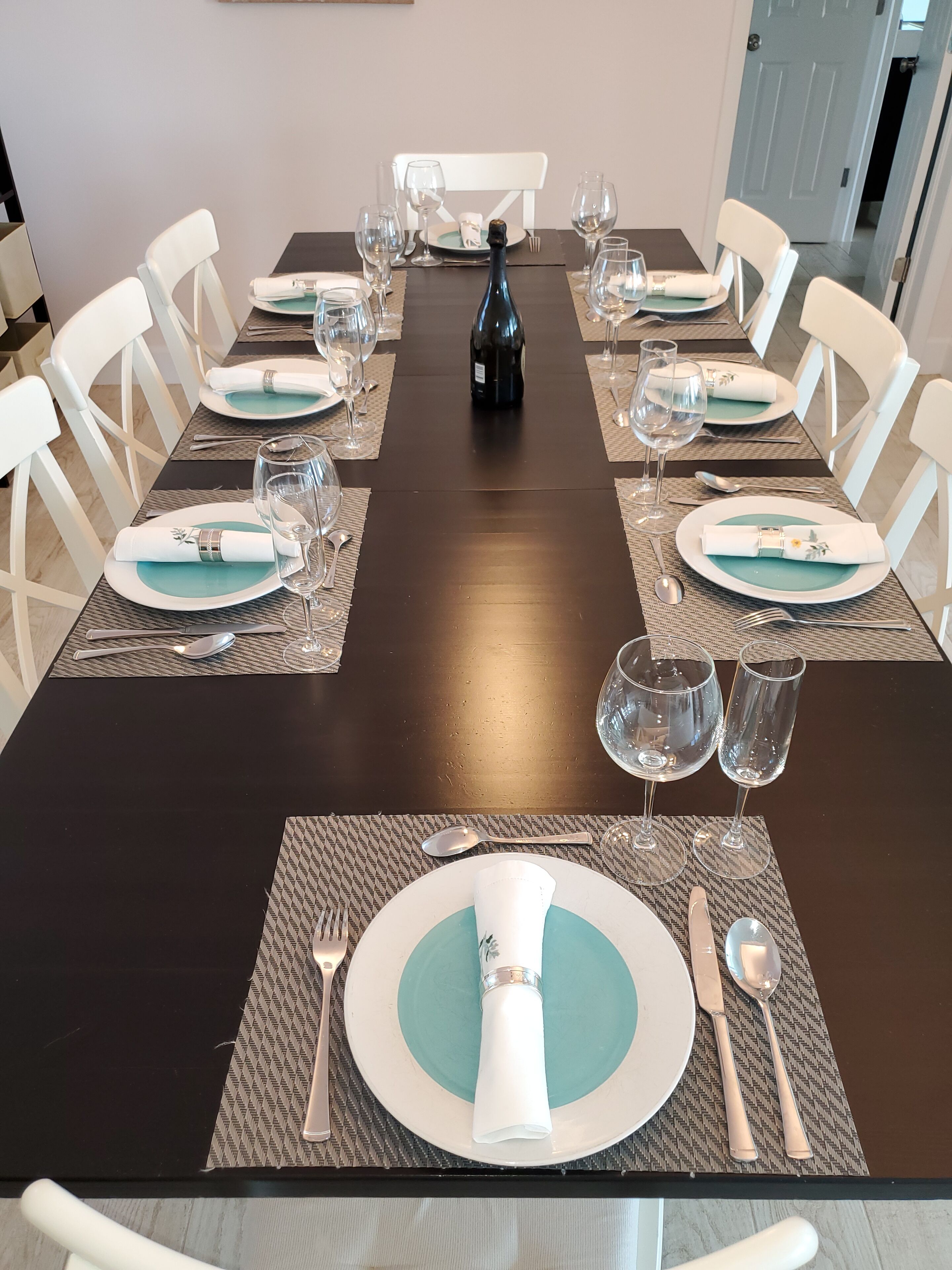 Une table à manger dressée pour six personnes, avec des chaises blanches, une bouteille de champagne et des verres à vin vides.