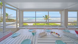 Un comedor con vista al océano, muebles blancos y una mesa dispuesta con platos, vasos y una cesta.