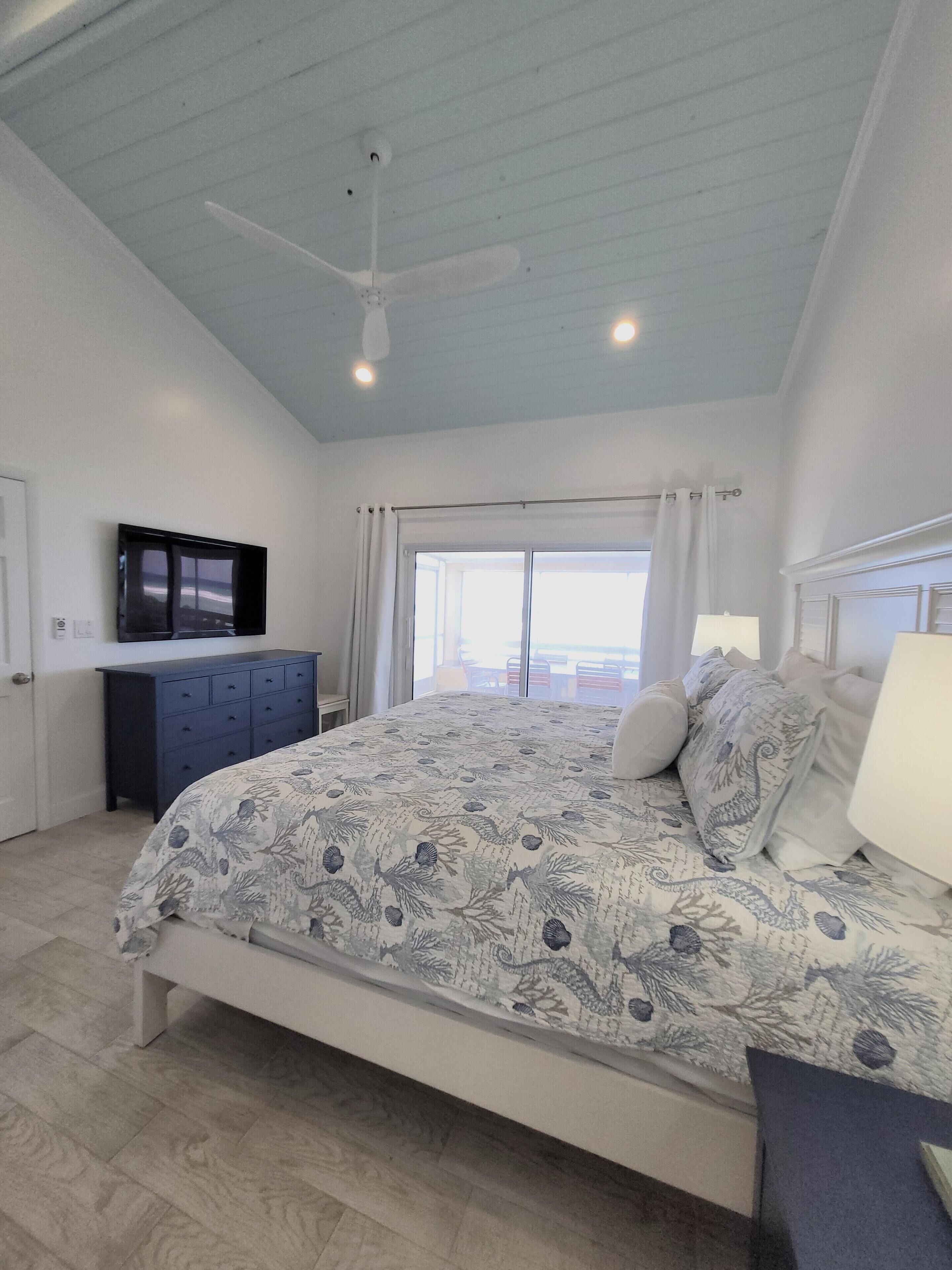 A bedroom with a bed, a ceiling fan, a TV, a window with curtains, and a view of the outside.