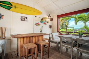 Uma área de bar de madeira com um prancha de surf, cadeiras e vista para o mar.