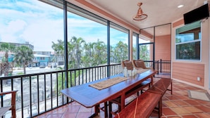 Varanda com mesa e bancos de madeira, janelas amplas com vista para palmeiras e edifícios, e um ventilador de teto.