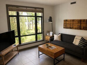 A living room with a sofa, coffee table, TV, and a large window with a view of greenery. - Beautiful 2nd Floor Condo on Quiet Creekside (Kelowna)