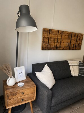 Una sala moderna con un sofá gris oscuro, una mesita de noche de madera con una lámpara, un cuadro enmarcado y una obra de arte en la pared.
