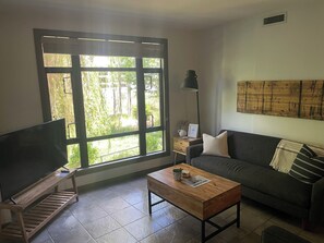 A living room with a large window, a flat-screen TV, a wooden coffee table, a sofa, and a floor lamp.