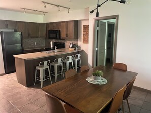 A modern kitchen with dark wooden cabinets, a black refrigerator, a microwave, and a sink. There is a dining area with a wooden table and four white bar stools.