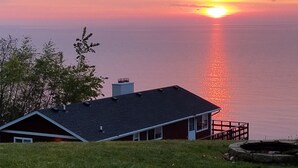 Een huis met een donker dak, schoorsteen en terras met uitzicht op een wateroppervlak tijdens de zonsondergang.