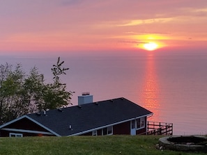 Une maison avec un toit sombre, une cheminée et une terrasse donnant sur une étendue d’eau au coucher du soleil.