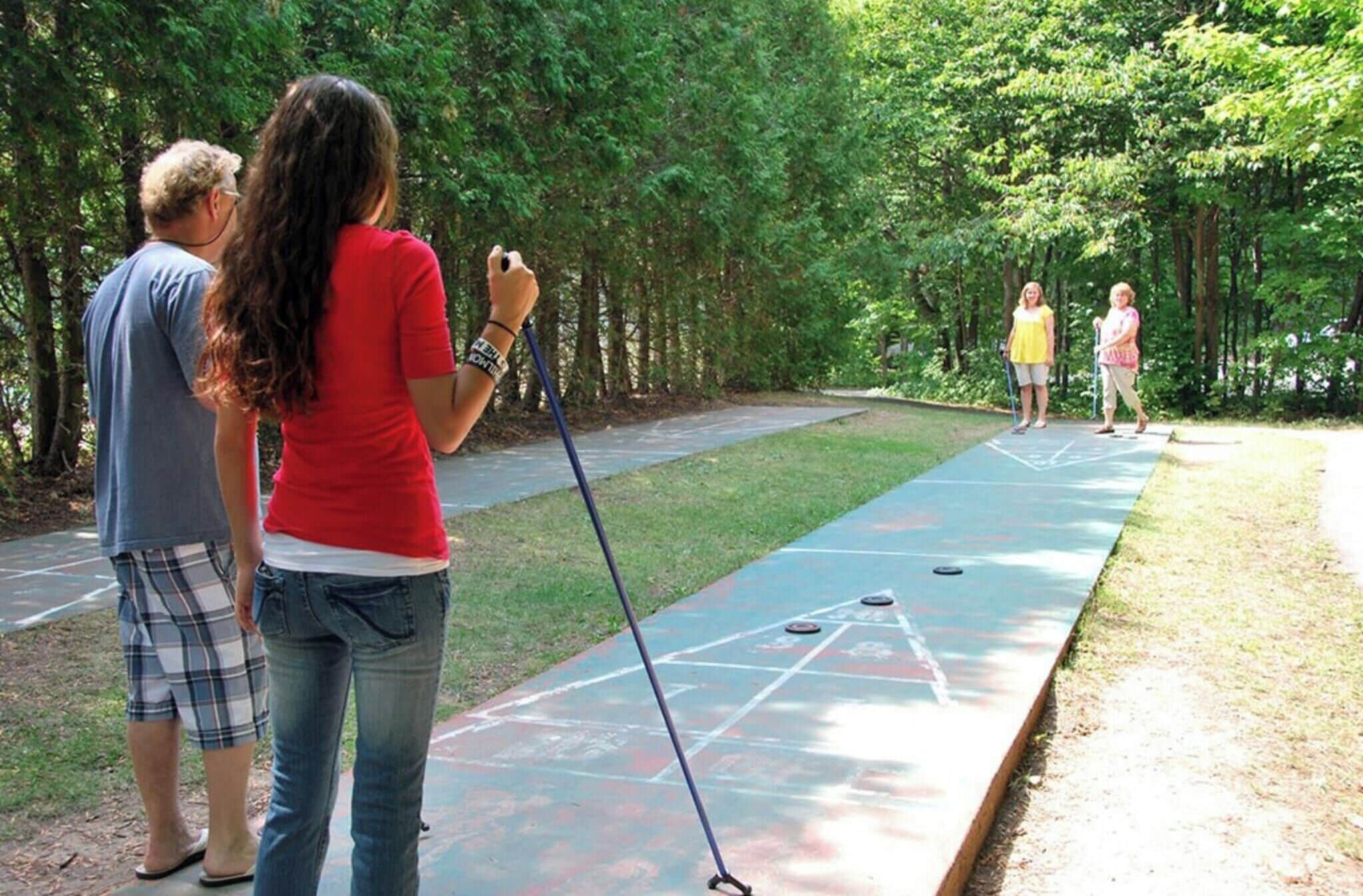 Un grupo de personas jugando al shuffleboard en una cancha verde al aire libre rodeada de árboles.