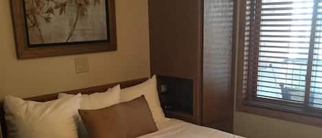 Una cama bien hecha con ropa de cama blanca y marrón, un cuadro con motivos florales encima y una ventana con persianas a la derecha.